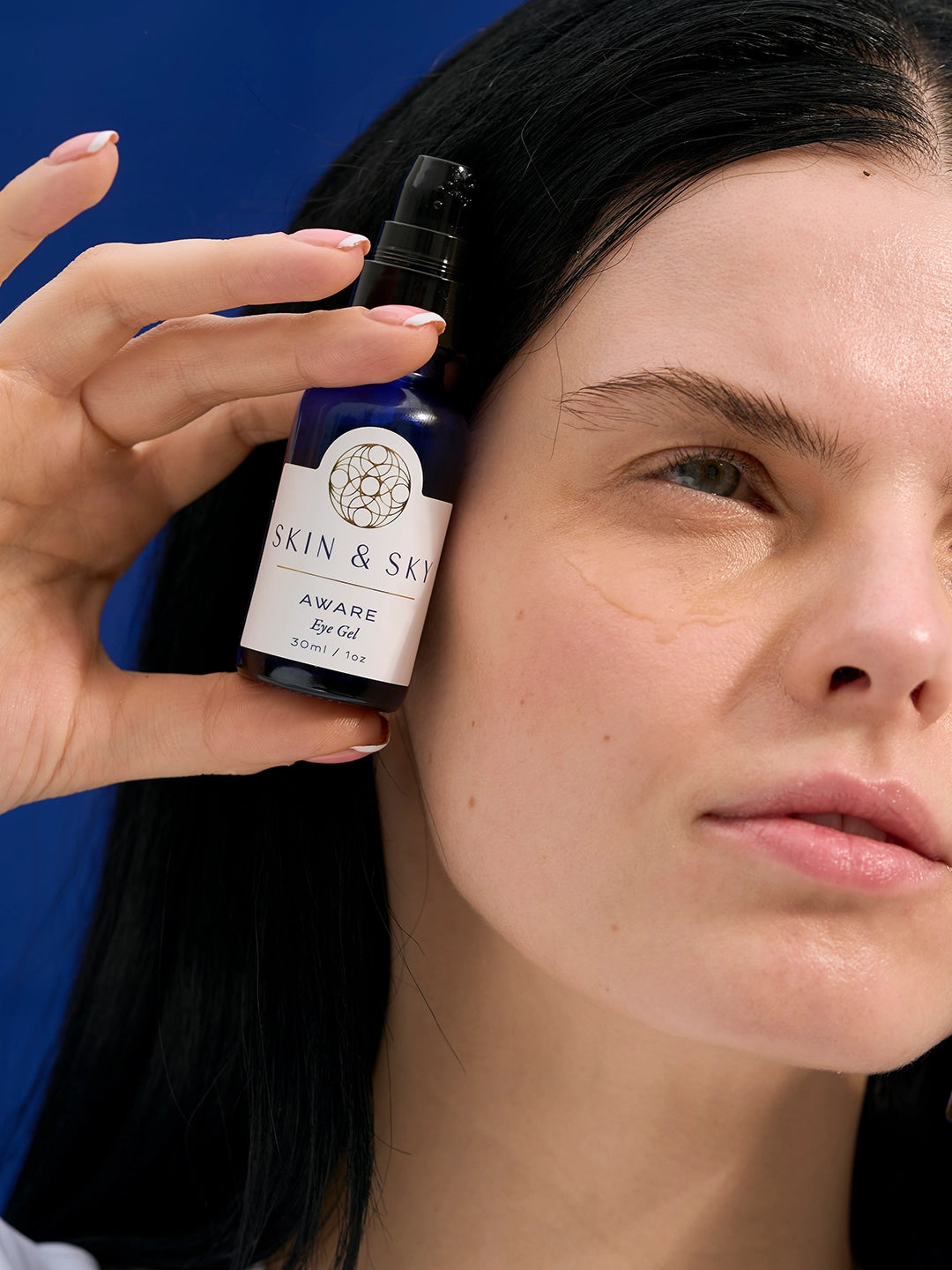 Una modelo sostiene el frasco de Aware Eye Gel junto a su rostro, mostrando una gota de gel transparente y ligero aplicado bajo el ojo para una hidratación inmediata.