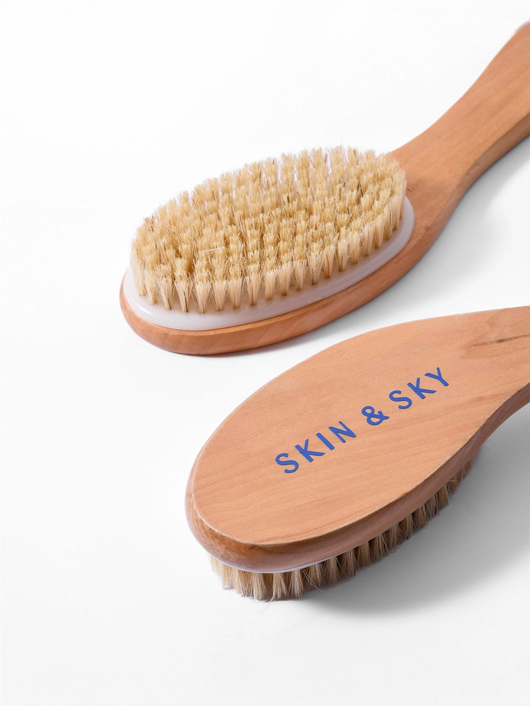 Primer plano de dos cepillos corporales Skin & Sky de madera; uno muestra las cerdas naturales densas para exfoliación y el otro destaca el logo de la marca grabado en azul sobre la madera pulida.