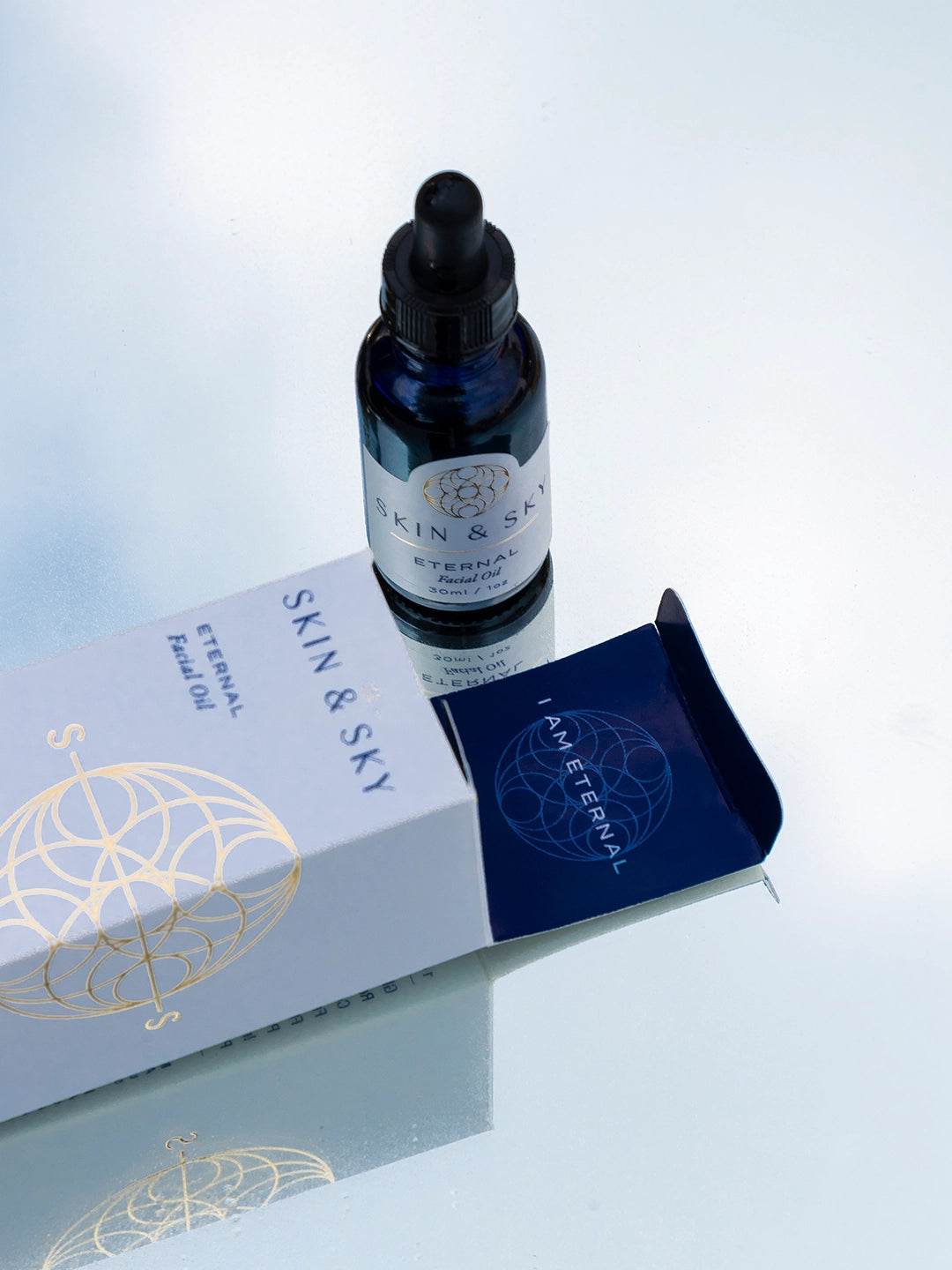 Botella de vidrio azul oscuro de Eternal Facial Oil de 30 ml / 1 oz junto a su caja premium blanca con detalles dorados y logo etéreo de Skin & Sky. Gotero negro y etiqueta elegante con diseño geométrico dorado. Aceite facial hidratante y reparador para todo tipo de piel, A.M. y P.M.