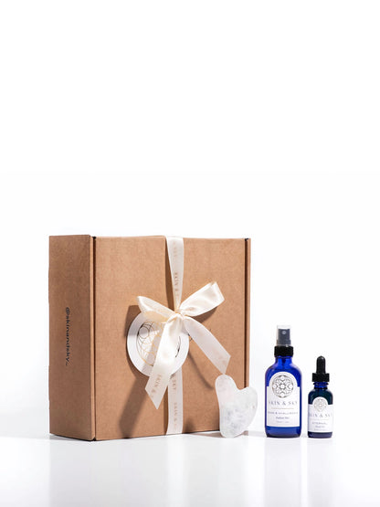 Presentación de regalo del Eternity Set de Skin & Sky: una caja de cartón kraft decorada con un lazo de satén blanco y etiqueta de la marca, junto al aceite facial Eternal, el tónico Rose & Hyaluronic y una Gua Sha de cuarzo blanco.