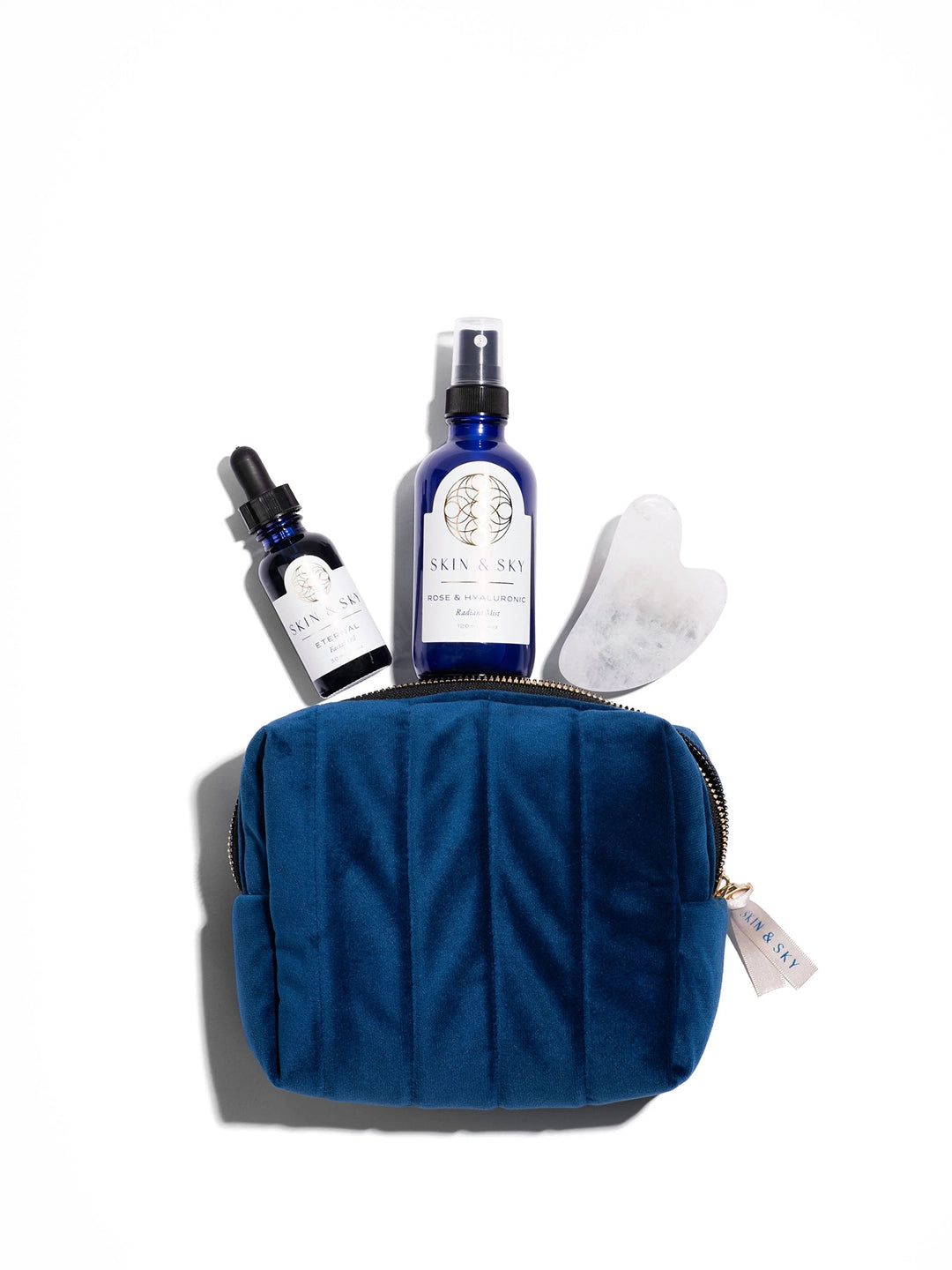 Vista superior de los elementos del Eternity Set: el aceite Eternal, el tónico Radiant Mist y la Gua Sha de cuarzo blanco AAA sobre una exclusiva cosmetiquera de terciopelo azul acolchada con el logo de Skin & Sky.