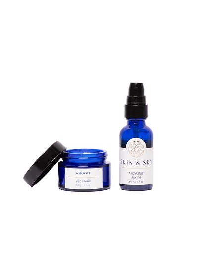 Modelo aplicando el Gaze Set de Skin & Sky: sostiene el AWARE Eye Gel y la AWAKE Eye Cream frente a un fondo azul. Se muestran puntos de producto bajo los ojos para ilustrar la rutina de hidratación y rejuvenecimiento de la mirada.