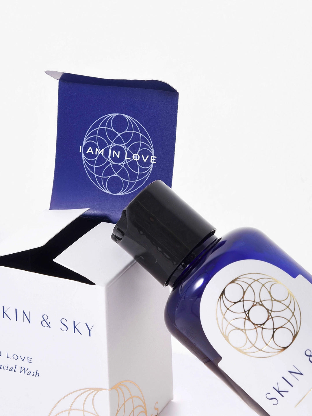 Frasco azul de In Love Facial Wash junto a su caja premium abierta con mensaje 'I AM IN LOVE' en blanco y geometría sagrada dorada. Producto de limpieza facial de Skin & Sky, 120 ml / 4 oz.