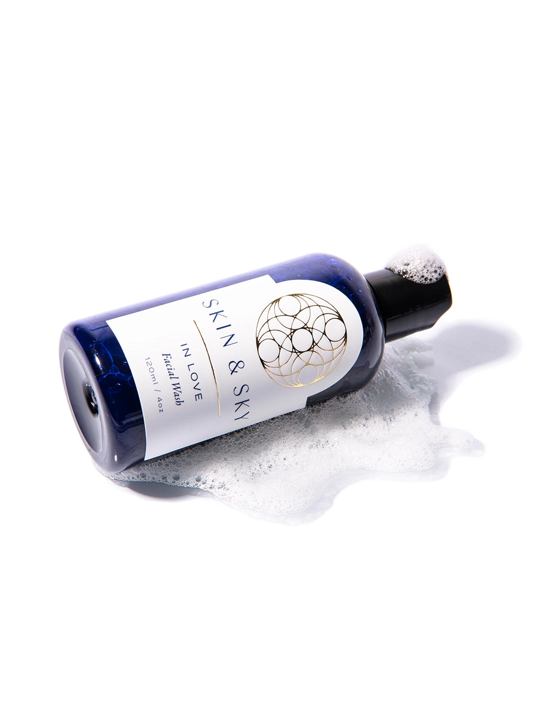 Botella azul de In Love Facial Wash acostada sobre espuma cremosa y burbujas blancas, mostrando su textura espumosa ligera y refrescante. Facial Wash de 120 ml / 4 oz que limpia suavemente, equilibra e hidrata la piel con ingredientes naturales como menta, eucalipto y miel, ideal para rutina A.M. y P.M.