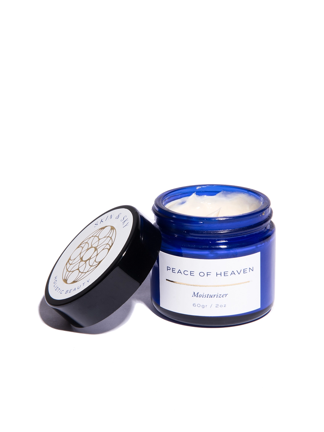 Primer plano del hidratante Peace of Heaven abierto, mostrando su textura cremosa y ligera de color blanco dentro de un envase de vidrio azul cobalto.
