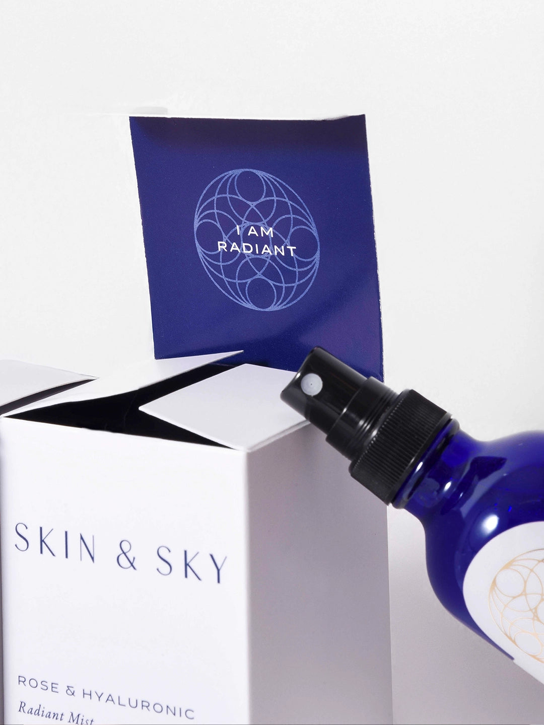 Detalle del empaque blanco de Skin & Sky abierto, revelando el mensaje "I AM RADIANT" en el interior y el atomizador negro de la bruma Rose & Hyaluronic Radiant Mist.
