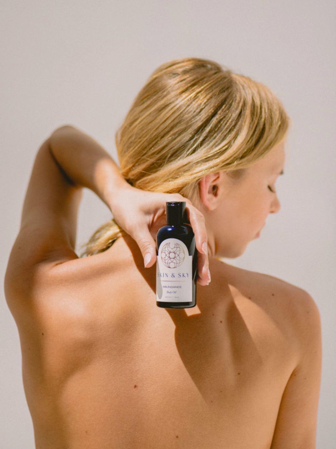 Una mujer sostiene la botella de Abundance Body Oil contra su espalda descubierta bajo la luz del sol, resaltando una piel hidratada y saludable.