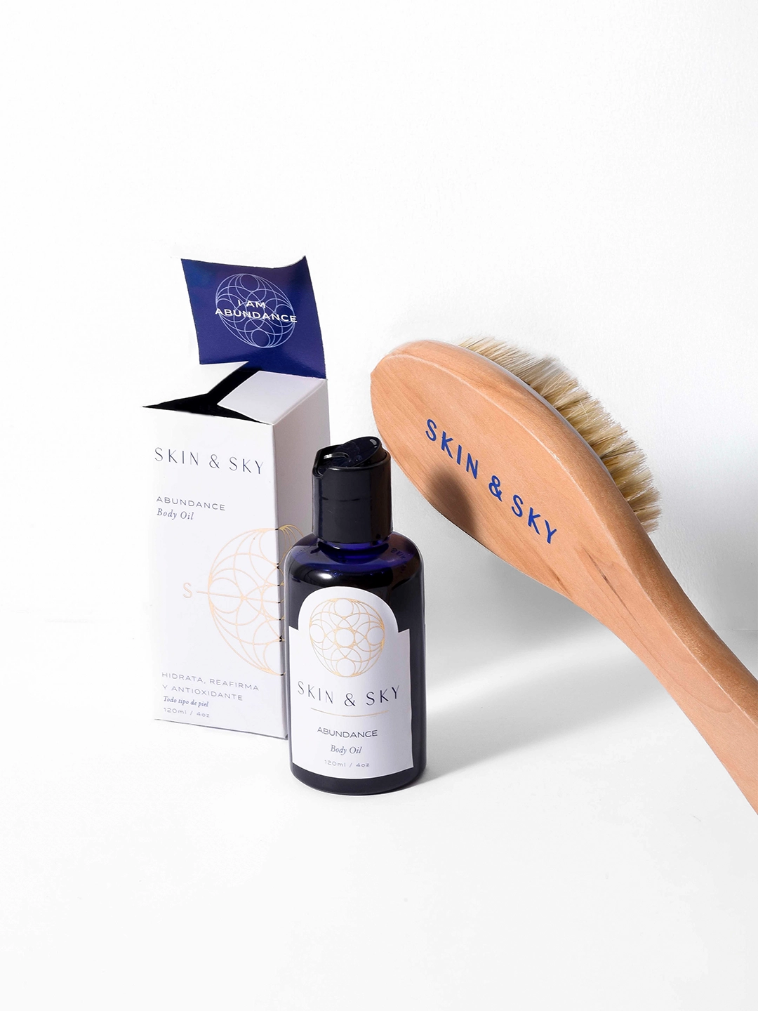 Botella de aceite corporal Abundance junto a su caja y el mango de un cepillo de madera Skin & Sky, presentados sobre un fondo blanco para una rutina de hidratación y rejuvenecimiento de la piel.