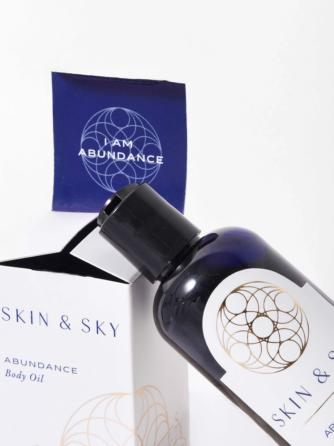 Primer plano del envase de Skin & Sky y su caja blanca; la solapa interior revela el mensaje "I AM ABUNDANCE” junto al frasco de aceite.