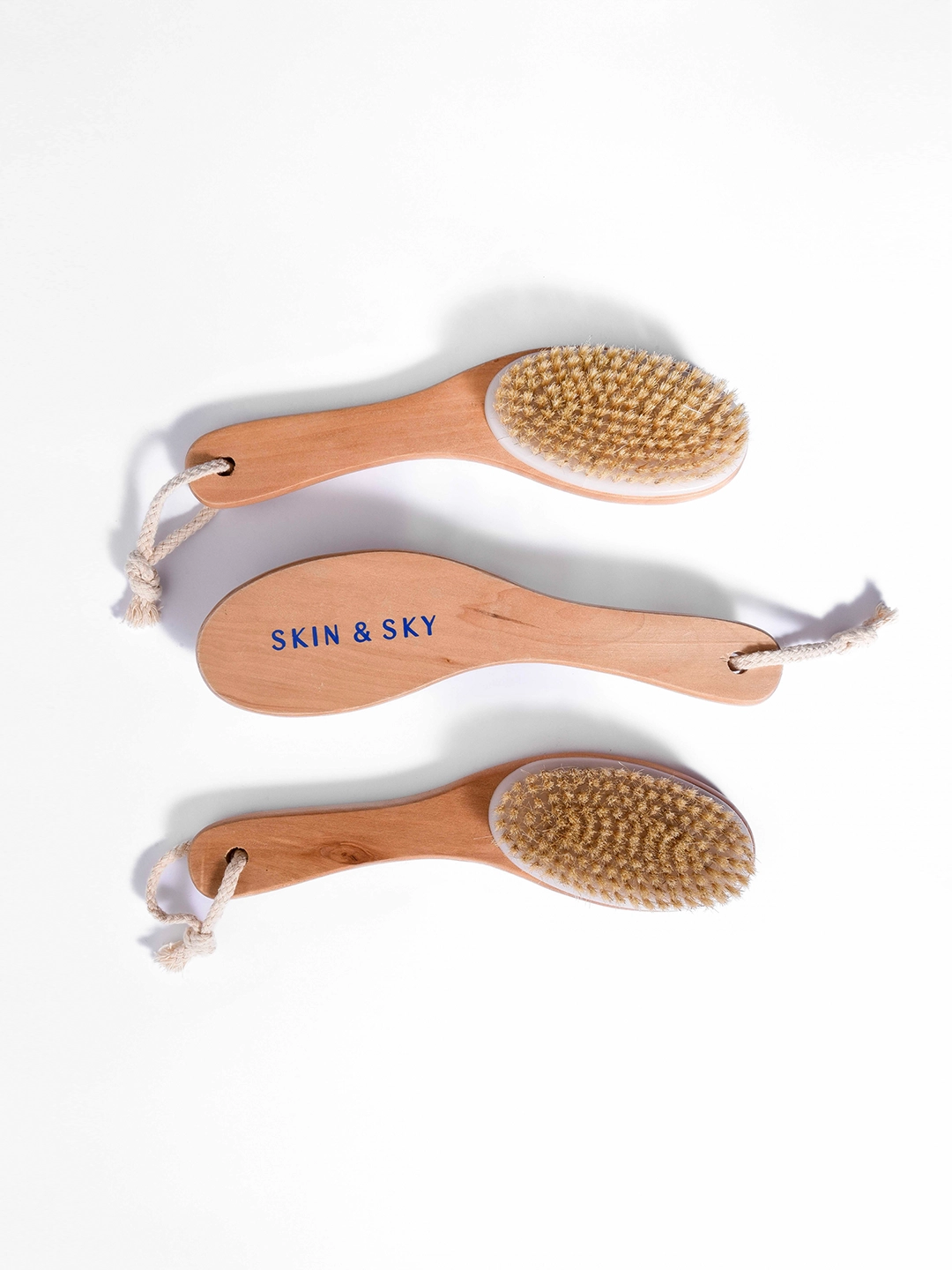 Vista aérea de tres cepillos corporales de madera con cerdas naturales y cordón de soga para colgar, dispuestos de forma organizada para mostrar el diseño ergonómico de Skin & Sky.