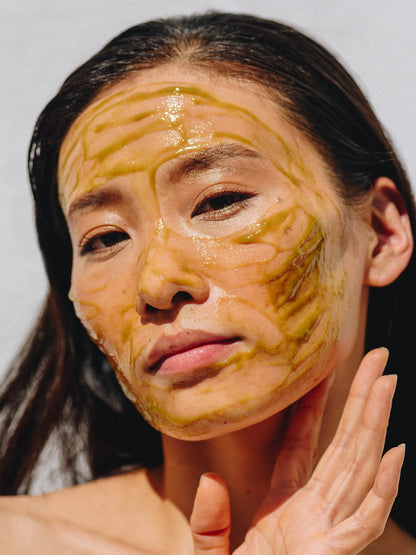 Mujer aplicando el Empower Stimulating Masque en el rostro, mostrando capas gruesas de mascarilla en tonos amarillo-verdoso brillante. Textura estimulante que rejuvenece, desintoxica y reafirma la piel con ingredientes naturales como algas y adaptógenos.