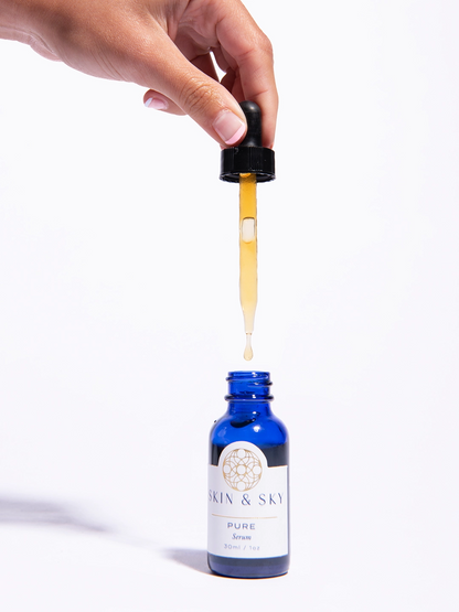 Una mano sostiene el gotero de Pure Serum de Skin & Sky extrayendo el suero de color ámbar de su envase azul, ideal para una aplicación precisa.