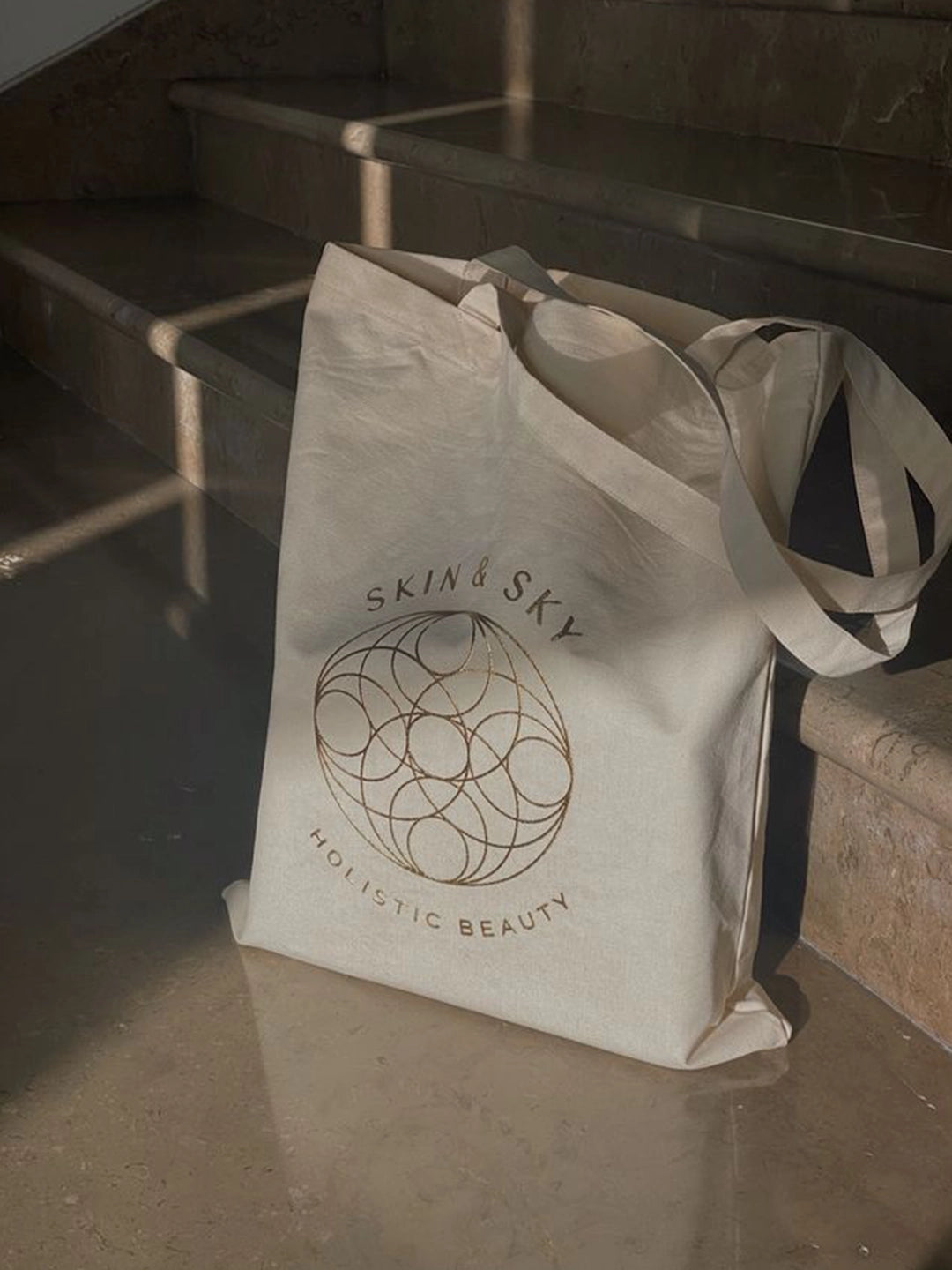 Beige tote bag with 'Skin & Sky' logo on a stone surface